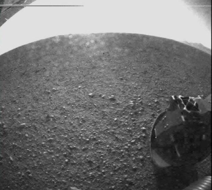 Třetí snímek z Marsu přijatý z Curiosity. Krytka navigační kamery už byla odklopena, vidíme tedy poměrně detailně rovnou pláň uvnitř kráteru Gale, jehož okraj je patrně v pozadí na snímku nahoře vpravo. NASA/JPL